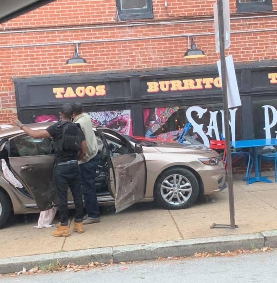 A damaged car resting on the sidewalk directly outside a restaurant's front windows and patio seating