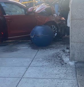 A red car driven into the corner of a hotel building, with bystanders and debris around it