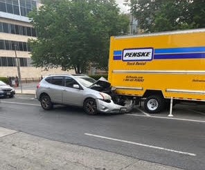 A silver SUV with a crushed front end stopped against the back of a parked yellow rental moving truck