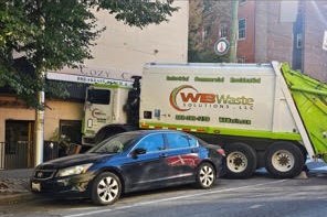 A trash truck stopped beside a parked blue car on a narrow residential street