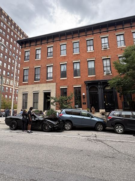 A black car with its front end crushed, collided into a blue SUV and a black SUV parked along a residential block, with bystanders on the sidewalk